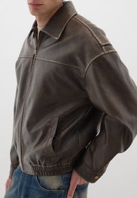 Giacca in pelle marrone con chiusura a zip, colletto a punta, vita elastica, tasche laterali, cuciture visibili e una finitura testurizzata.