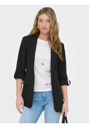 Kobieta o długich blond włosach ubrana w czarną marynarkę, białą koszulkę z małymi wzorami kokardek oraz jasnoniebieskie dżinsy, trzymająca czarną torebkę.