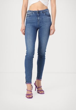 Frau trägt blaue Skinny-Jeans mit mittlerem Bund und Reißverschlüssen am Knöchel sowie lila Sandalen mit hohen Absätzen und steht vor weißem Hintergrund.