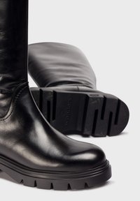 Botas de cuero negro hasta la rodilla con punta redonda, superficie texturizada y suela de goma negra resistente con ranuras horizontales.