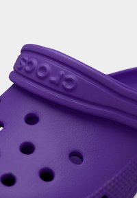 Κοντινή λήψη του ιμάντα παπουτσιού Crocs σε μωβ χρώμα με οπές αερισμού και υφή στην επιφάνεια, που δείχνει το χαραγμένο λογότυπο "CROCS" στον ιμάντα.