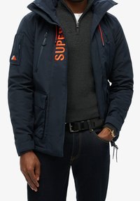 Homme portant une veste bleu marine avec des détails de fermeture éclair orange sur un pull gris foncé zippé et un jean foncé, une main dans la poche.