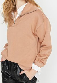 Mujer vistiendo un sudadera beige de media cremallera sobre una camisa blanca de botones, combinada con pantalones de imitación cuero negros, con las manos en los bolsillos.
