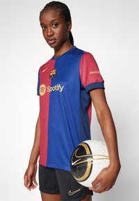 FC Barcelonan pelipaita on sinipunainen kultaisin yksityiskohdin, jossa on logo ja "Spotify"-teksti; yhdistetty mustiin shortseihin ja kuvioituun jalkapalloon.