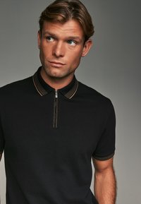 Polo shirt noir avec un col zippé et des accents rayés dorés, doté d'un tissu texturé et de manches courtes.