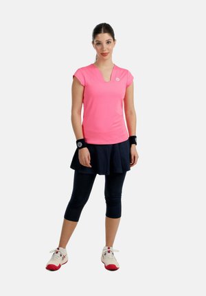 Vrouw die staat en een roze top met korte mouwen draagt, een zwarte rok over korte legging, zwarte polsbandjes en witte sportschoenen met rode zolen.