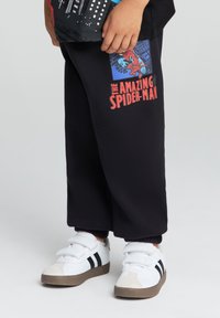 Pantaloni della tuta neri con una grafica di Spiderman in blu e rosso. Abbinati a sneaker bianche, con strisce nere e suola marrone.