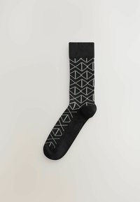 Chaussette noire avec des motifs géométriques blancs, comprenant des losanges et des triangles. Fabriquée en matériau doux avec un bord et un orteil noirs solides.
