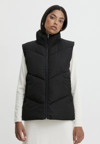 Gilet rembourré noir avec un col montant, fermeture éclair et motif en chevrons. Fabriqué en tissu léger, il présente une texture lisse et un design sans manches.