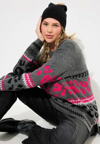 Maglione grigio lavorato a maglia con motivi leopardati rosa e geometrici sulle maniche, abbinato a jeans neri e un berretto nero. Tessuto texturizzato.