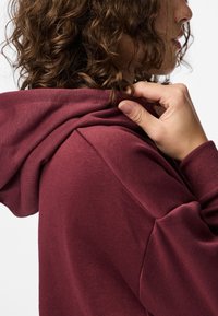 Persona con cabello rizado castaño ajustando la capucha de un sudadera color burdeos, mostrada de lado y por detrás sobre un fondo blanco.