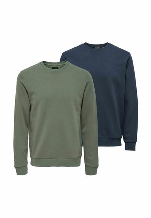 Junger Mann, der einen dunkelblauen Pullover, blaue Jeans und braune Schuhe trägt, vor einem schlichten hellen Hintergrund steht.
