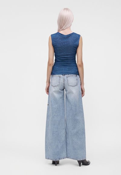 Femme aux longs cheveux clairs portant un haut bleu sans manches, un jean bleu clair à jambes larges et des chaussures noires à talons, debout de dos.