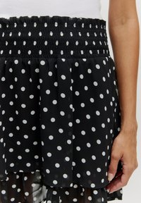 Zwarte polka dot rok van lichtgewicht stof, met een smocktail en gelaagde ruffles met witte cirkelpatronen erdoorheen.