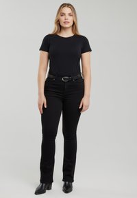 Vrouw staand, naar voren kijkend, gekleed in een zwart T-shirt, zwarte spijkerbroek, zwarte riem en zwarte enkellaarsjes met hak, tegen een effen grijze achtergrond.