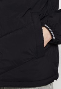 Giubbotto imbottito nero con tasca con zip, polsini elastici e accentuazione del logo sulla manica. Tessuto liscio con dettagli di cucitura minimi.