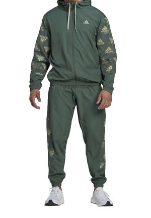 Mężczyzna ubrany w zielony dres marki Adidas z kapturem i zapinana na zamek kurtką oraz spodniami ozdobionymi wzorami z logo na rękawach i bokach, w komplecie z białymi trampkami.