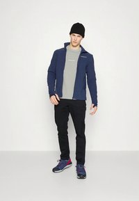 Tumši zils flīsa žakete, pelēks t-krekls, melnas bikses un zilas kurpes ar sarkaniem akcentiem. Vīrietis stāv ar vienu roku paceltu, fonā ir vienkāršs fons.