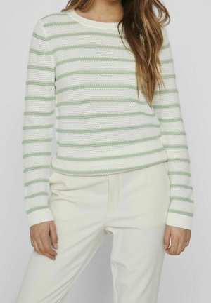 Mujer que lleva un jersey blanco con rayas horizontales verde claro y pantalones blancos, de pie con las manos relajadas a los lados.