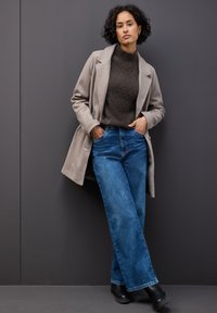 Cappotto in misto lana beige con colletto oversize, maglione grigio scuro e jeans in denim blu a gamba larga. Stivaletti neri alla caviglia completano il look.