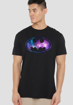 Mann trägt schwarzes T-Shirt mit buntem Galaxy-Batman-Logo, schaut seitlich vor einem hellen, einfarbigen Hintergrund.