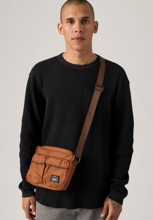 Bărbat îmbrăcat cu un pulover texturat negru și pantaloni gri, având o geantă crossbody maro cu două buzunare frontale și o emblemă.