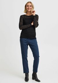 Zwarte kanten top met lange mouwen en een gestructureerd patroon, gecombineerd met marineblauwe taillepantalons en zwarte hakken enkellaarsjes. Gemodelleerde, eigentijdse silhoët.