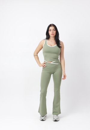 Joven mujer de pie con camiseta sin mangas verde claro y pantalones acampanados a juego, con zapatillas blancas sobre fondo blanco.