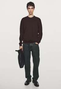 Dunkelbrauner Pullover, grüne Jeans, schwarze Lederschuhe, hält eine schwarze Ledertasche. Schlichtes Design, strukturierter Stoff, lockere Passform und standardmäßige Details.