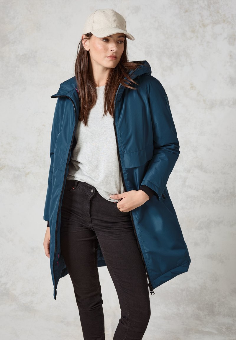 Lång, huva jacka i mörk teal, tillverkad av ett glänsande, slätt material. Bärs över en ljusgrå skjorta och svarta jeans. Anmärkningsvärd detalj: dragkedja fram.