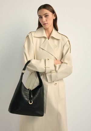 Kobieta w beżowym płaszczu trencz, trzymająca dużą czarną, teksturowaną skórzaną torebkę na ramię z złotym pierścieniem na tle jednolitym.
