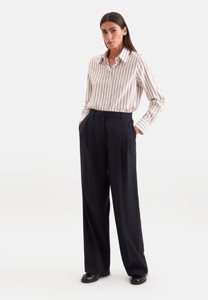 Kvinde med langt mørkt hår iført en beige og hvid striped knap-op skjorte, der er stoppet ned i sorte bukser med vide ben, med hænderne i lommerne.