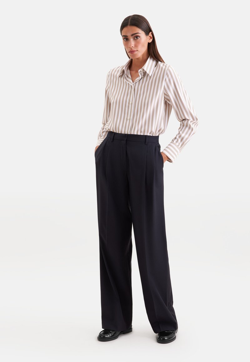 Femme aux longs cheveux foncés portant une chemise beige et blanche rayée boutonnée, rentrée dans un pantalon noir à jambes larges, les mains dans les poches.
