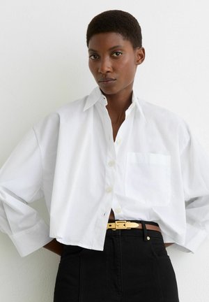 Lühikeste juustega naine, kes kannab lahtist valget lühendatud nööbiga särke ja musta kõrge vöökohaga pükse pruunika vööga, taustaks tavaline taust.