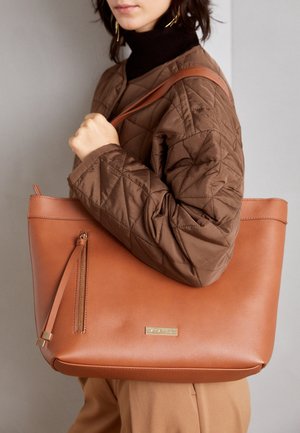 Vrouw draagt een bruine doorgestikte jas en een zwarte coltrui, en draagt een grote beige leren tas met een voorvak met rits.