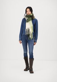 Bufanda de lana en tonos verde y blanco, llevada alrededor del cuello. La modelo viste una chaqueta de mezclilla azul, jeans ajustados y botas marrones hasta la rodilla.