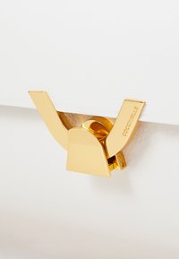 Il fermaglio in metallo dorato presenta una forma a V distintiva, con una superficie liscia e lucida. È fissato a un materiale bianco e testurizzato.