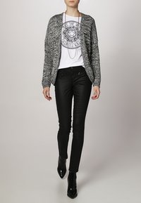 Černobílý vzorovaný kardigan přes bílou košili, kombinovaný s černými skinny džíny a černými špičatými kotníkovými botami. Viditelný řetízkový náhrdelník.