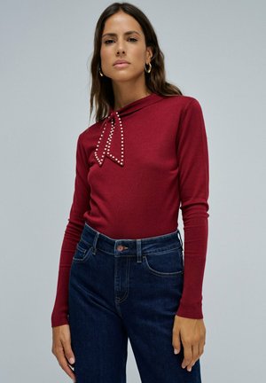 Mujer con una blusa roja de manga larga con detalle de moño incrustado de perlas y jeans de tiro alto de color azul oscuro, de pie contra un fondo liso.