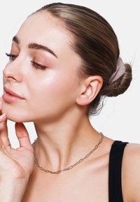 Scrunchie marrone nei capelli, collana di catena argentata, pelle liscia, trucco leggero, luce naturale, top nero, mano che tocca delicatamente il mento.