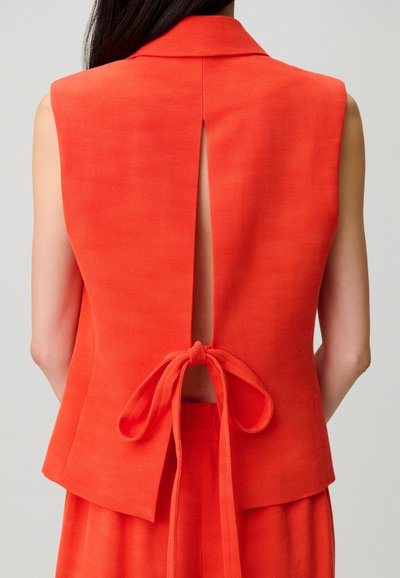 Top senza maniche di un arancione brillante con uno spacco sul retro legato a fiocco in vita, indossato da una persona con capelli scuri su uno sfondo neutro.