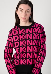 Megztinis su drąsiu rožiniu ir juodu geometriniu raštu su "DKNY" užrašu. Juodas banguotas apykaklė ir rankogaliai.