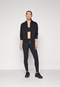 Giacca atletica nera con cappuccio, abbinata a un reggiseno sportivo nero e leggings aderenti. Il modello indossa scarpe da ginnastica nere. Tessuto liscio e elasticizzato.