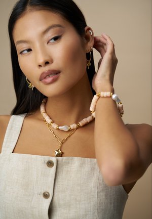 Perlen- und goldfarbener Schmuck mit pinkfarbenen Perlen, Sternanhänger und Kettendetail. Getragen zu einem beigen Leinenoutfit mit Knöpfen.