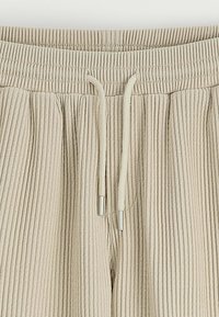 Pantalons de jogging beige à côtes avec une taille élastique, des cordons de serrage et un tissu texturé. Présente des accents métalliques argentés aux extrémités des cordons.