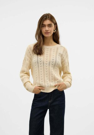 Maglione in crema con un motivo a lavorazione aperta, colletto tondo e polsini a costine, abbinato a jeans scuri. Tessuto morbido, vestibilità rilassata.