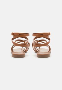 Sandalia de cuero marrón con múltiples correas entrelazadas, hebilla en el tobillo, suela plana y diseño minimalista que resalta una textura suave.