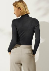 Frau mit kurzem blonden Haar, die einen engen schwarzen Langarmrollkragenpullover und beigefarbene hochtaillierte Hosen trägt, mit den Händen in den Taschen, abgewandt.