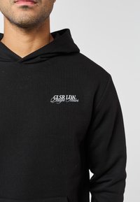 Mannelijke torso met een zwarte hoodie met witte geborduurde tekst "GLSR LDN Design Atelier" op de linkerborst tegen een effen achtergrond.