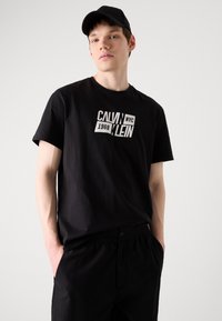 Fekete pamut póló rövid ujjal és kerek nyakkivágással, a "Calvin Klein NYC 1968" fehér grafikájú logójával. Fekete nadrággal párosítva.